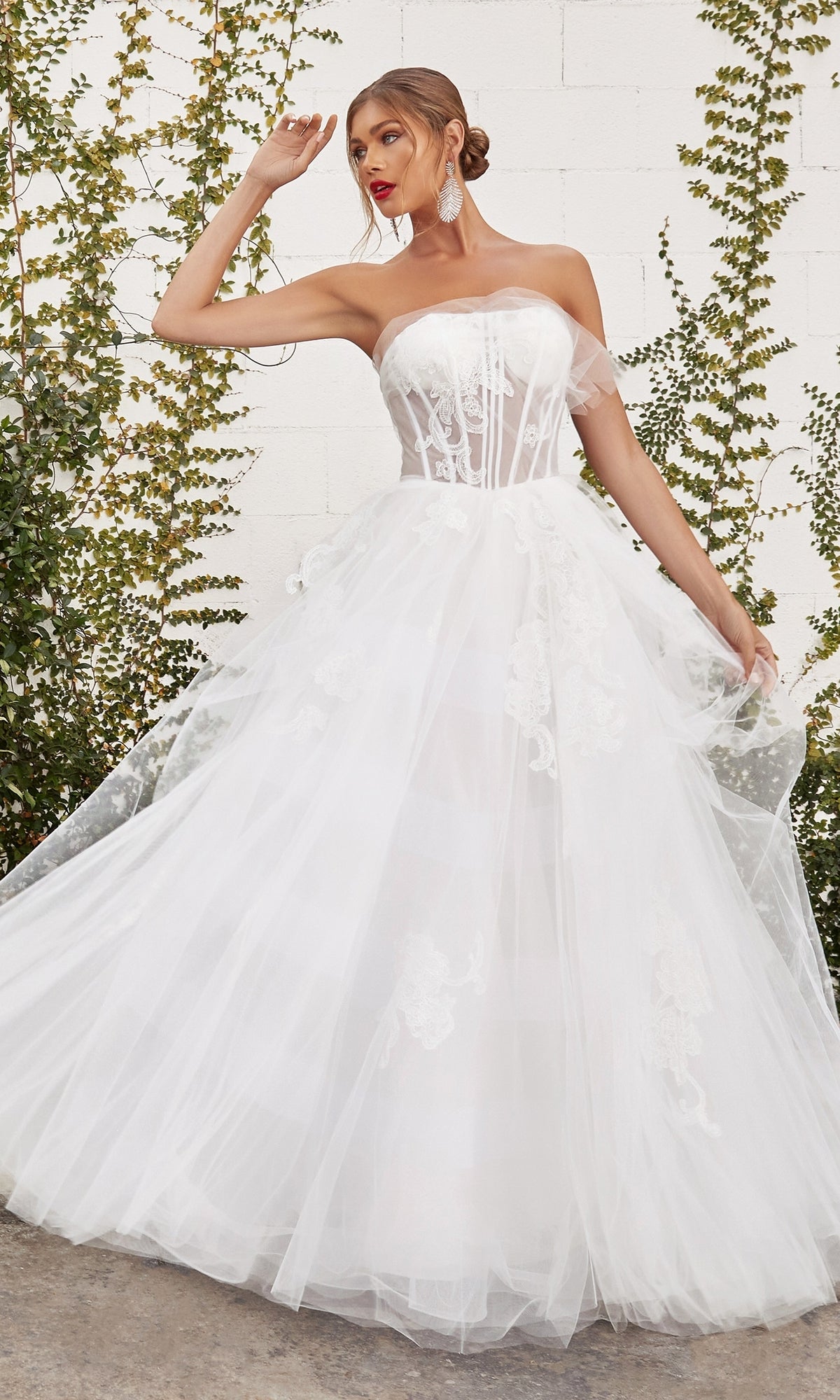 A woman stands outdoors against a white wall and greenery wearing the Andrea and Leo A1050W, a white strapless ball gown with a sheer corset bodice and floral lace details.